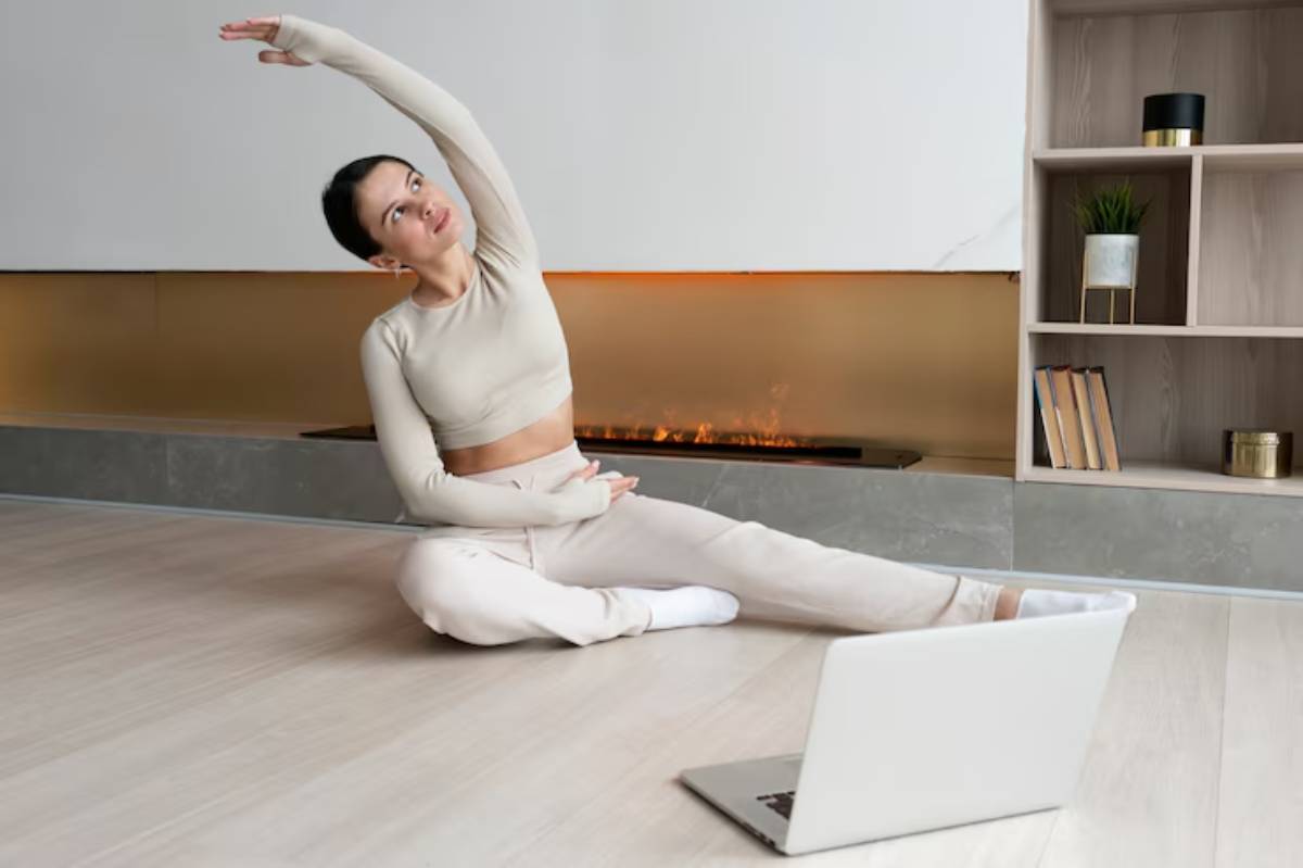 A person stretches their body to the side during a home workout session, with a laptop positioned in front and a cozy fireplace backdrop.