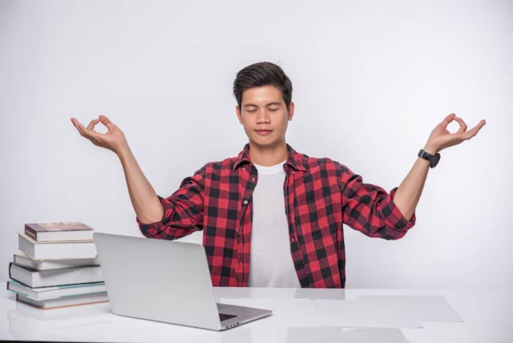 A person in a red checkered shirt sits at a desk with a laptop and books, gesturing in a meditative pose against a white backdrop.