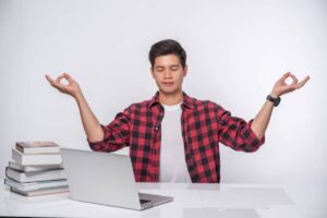 A person in a red checkered shirt sits at a desk with a laptop and books, gesturing in a meditative pose against a white backdrop.