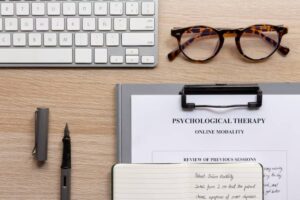 A workspace with a keyboard, tortoiseshell glasses, a notebook, and documents about online psychological therapy.