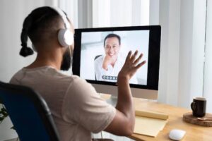 A person with headphones gestures during a video call, seated at a desk with a coffee mug and a notepad in a well-lit room.