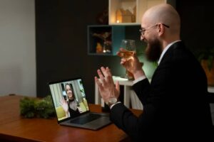 A man in a suit sits at a wooden table, toasting with a drink while video chatting on a laptop. Festive greenery is nearby.