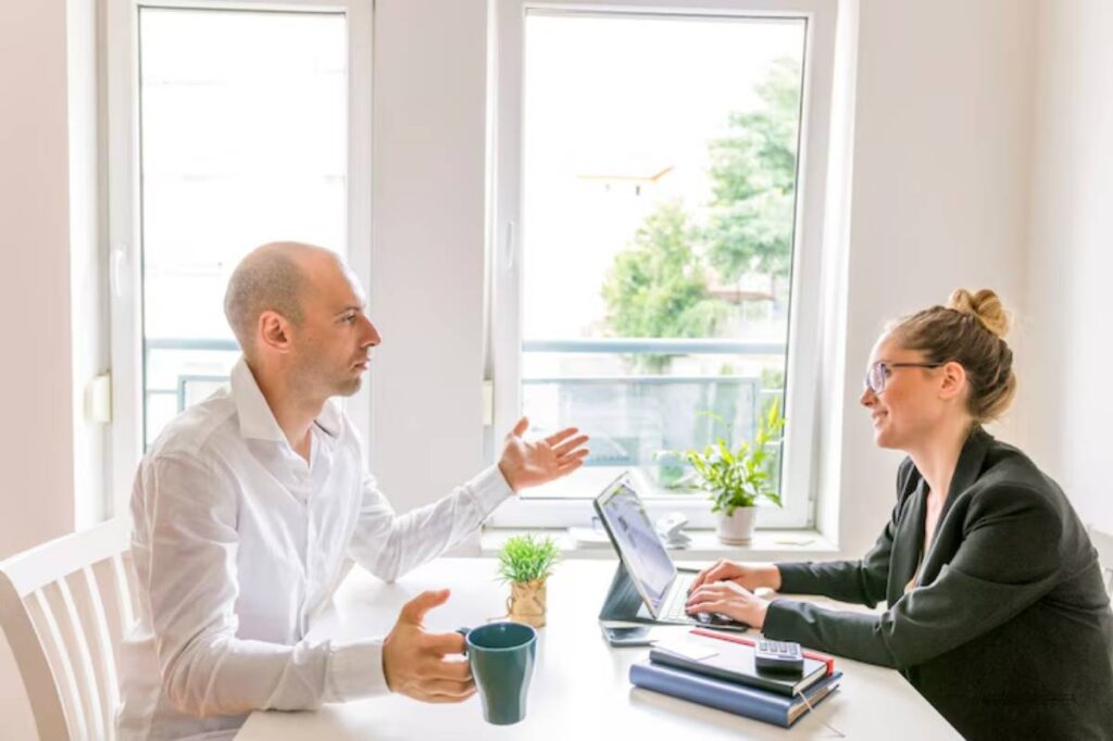 A man in a white shirt gestures while a woman in a black blazer types on a laptop at a bright, modern office table.