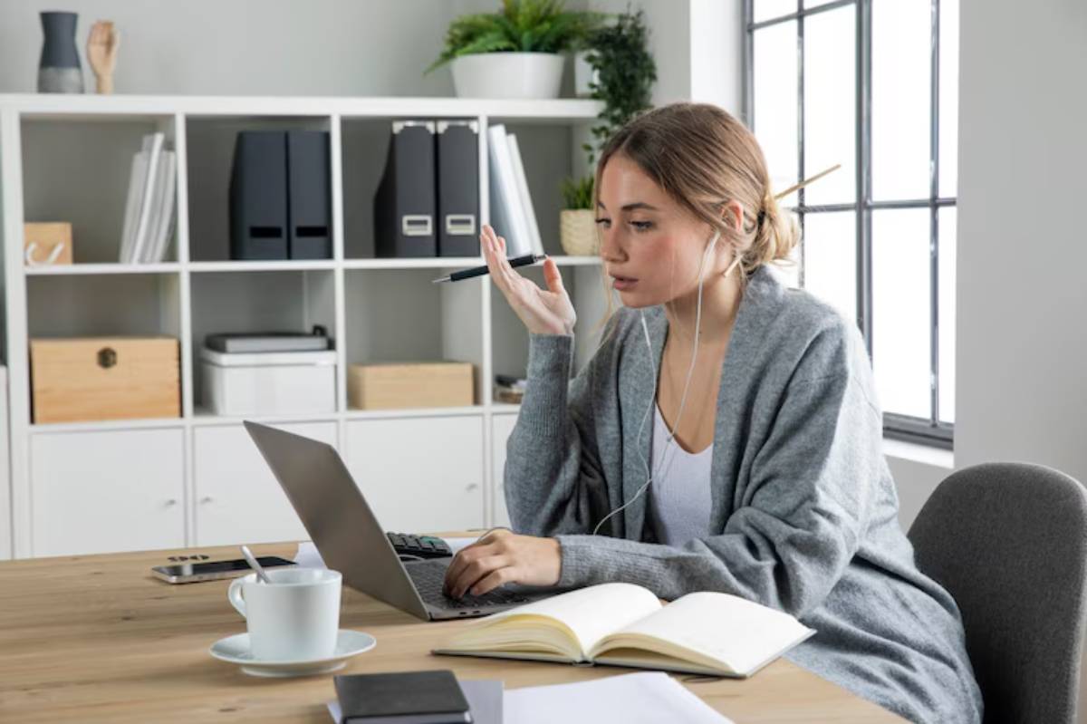  A woman in a cozy grey cardigan engages in a virtual conversation at a desk with a laptop, notebook, and a cup of coffee.