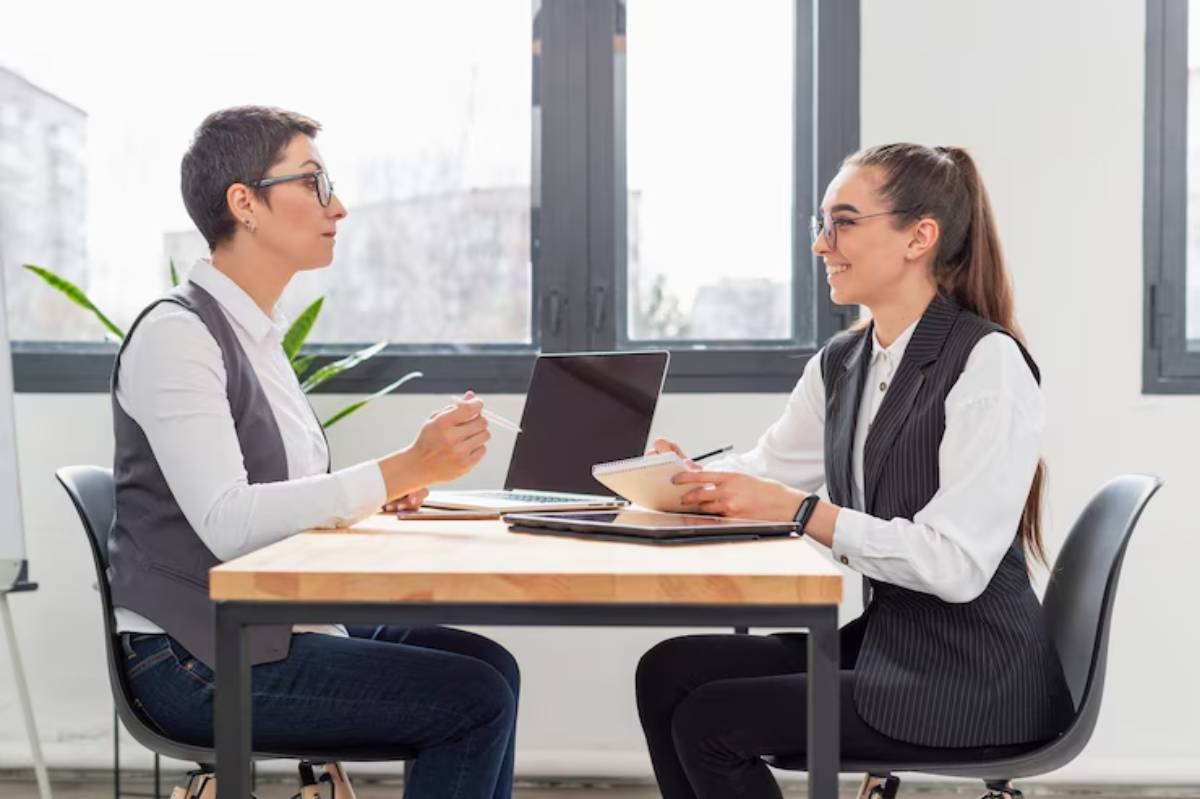 Two professionals engage in discussion at a wooden table, with a laptop and notepad in a bright, modern office space. 