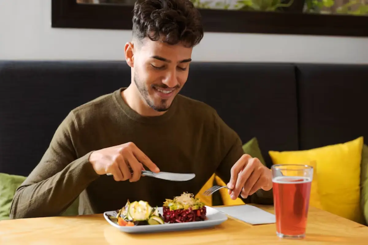 A person sits at a wooden table, poised to eat a colorful plate of salad and vegetables, with a glass of red beverage nearby. 
