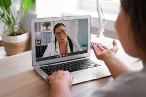 A person engaged in a video call on a laptop, gesturing with one hand, with indoor plants and a desk in the background.
