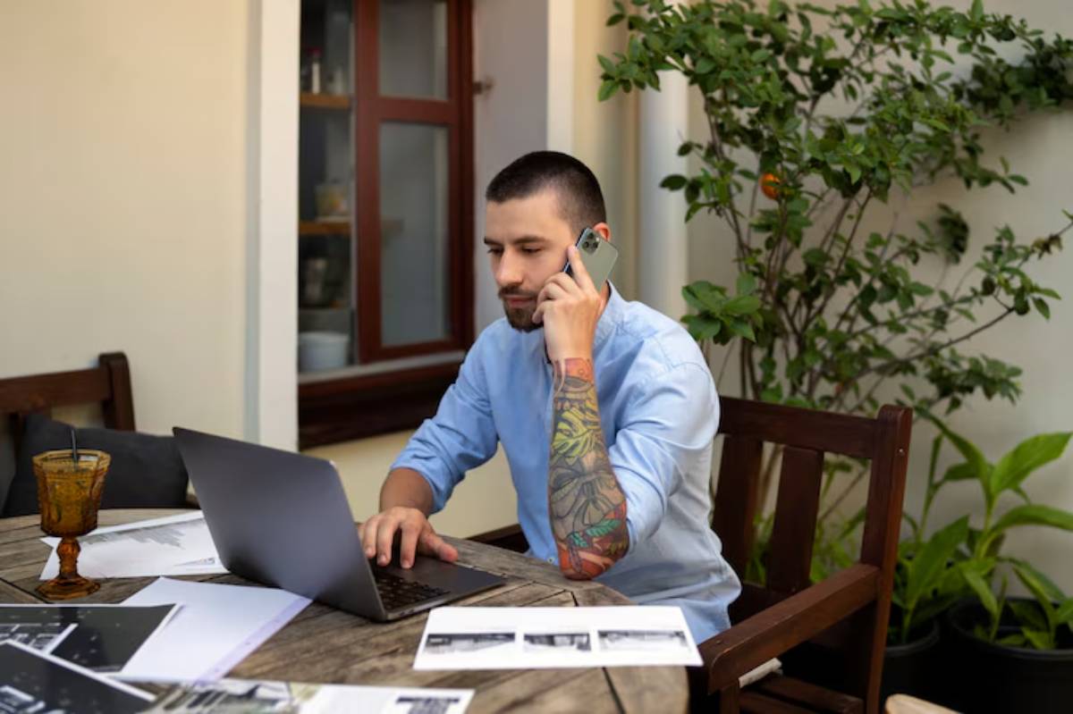  A person in a blue shirt is sitting at a wooden table with a laptop, surrounded by plants and papers, while on a phone call.
