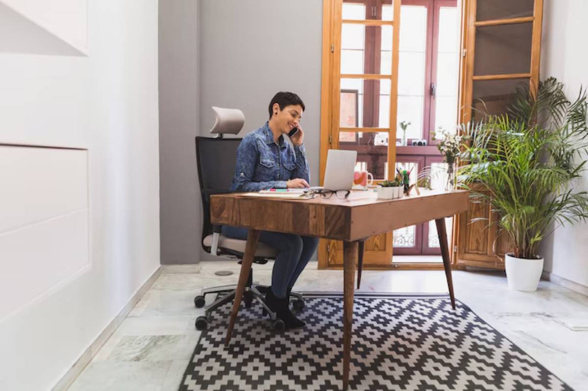 A person sits at a wooden desk with a laptop, surrounded by plants and a patterned rug, while on a phone call in a bright office. 