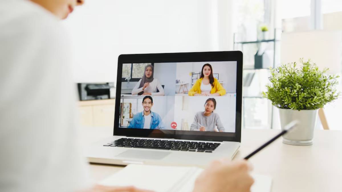 A person attends a video call on a laptop, showing multiple participants in a bright, modern workspace with a potted plant. 