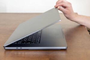 A hand lifts the lid of a sleek, silver laptop resting on a wooden table, revealing the keyboard and trackpad underneath.