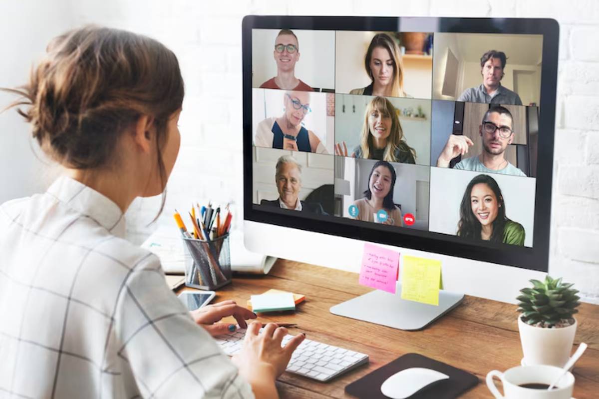 A person works at a computer in a cozy home office, participating in a video conference with multiple remote attendees visible on the screen. 