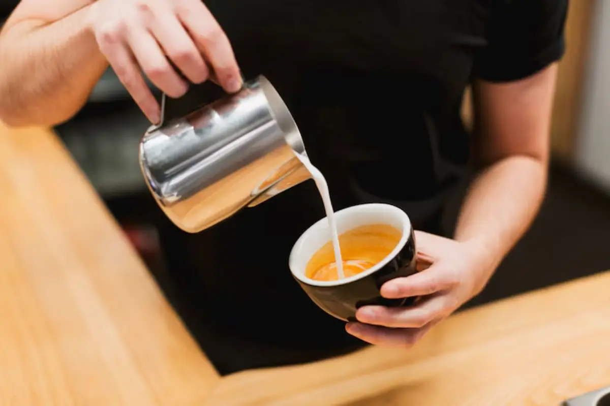A barista pours steamed milk from a metal jug into a black cup containing vibrant orange espresso, showcasing latte art in progress. 