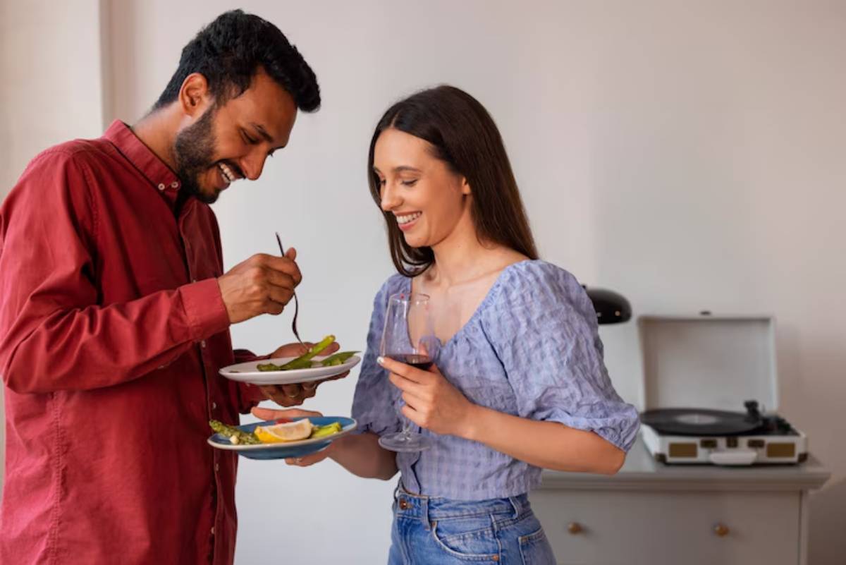  A man and a woman standing in a kitchen, each holding plates of food. They are smiling and wearing casual clothing, with kitchen appliances visible in the background.