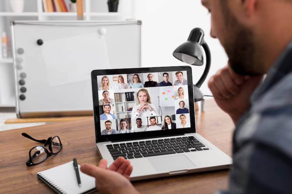  A person sits at a desk, engaged in a video conference on a laptop displaying multiple participants in a virtual meeting.