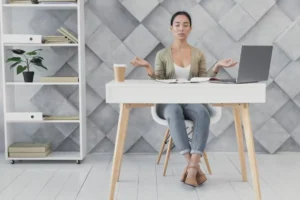 A person sits at a modern desk in a stylish office, practicing mindfulness, with a laptop and coffee cup nearby, surrounded by bookshelves.