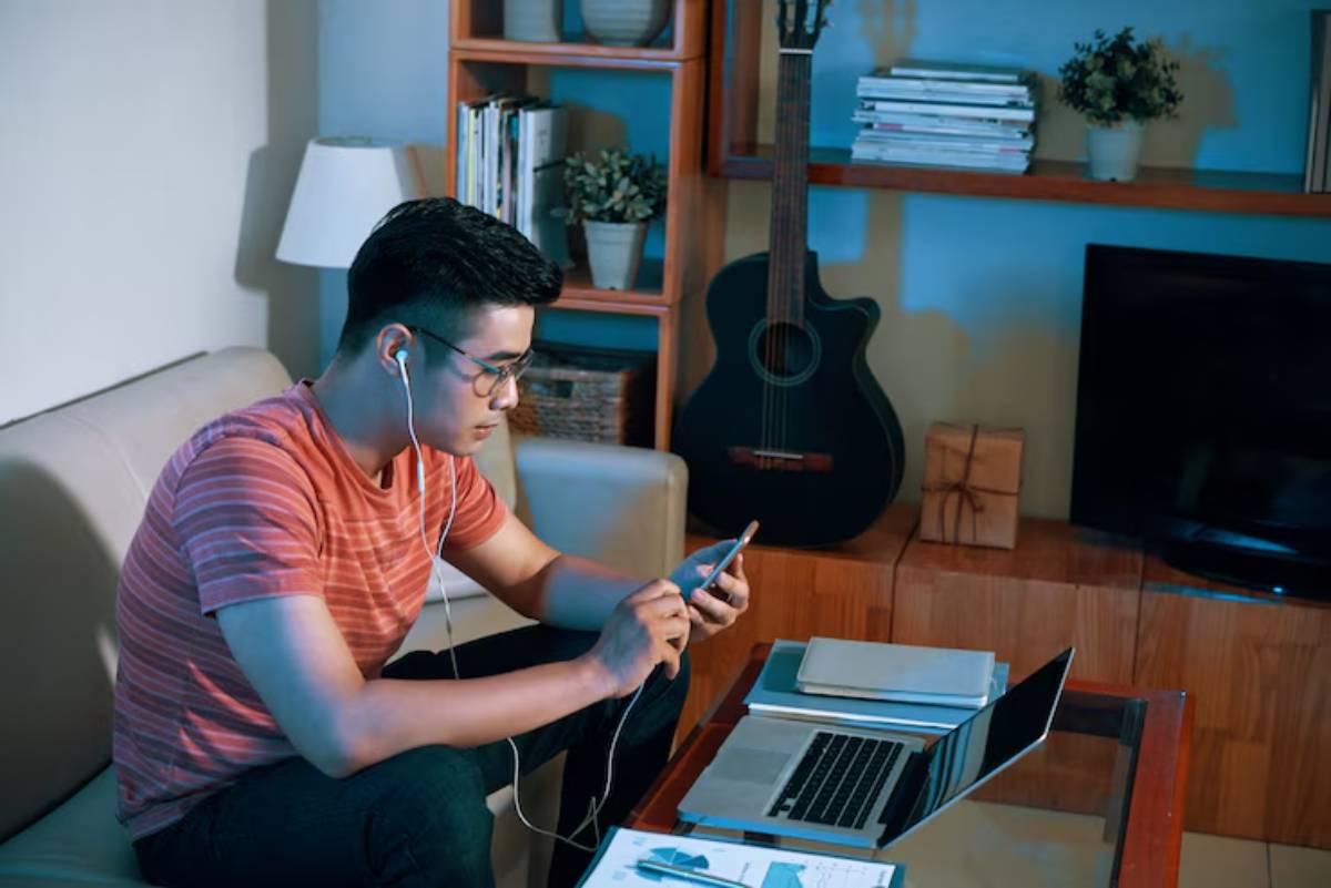 A person with earphones sits on a couch, using a smartphone, with a laptop and documents on a glass table in a cozy living room. 