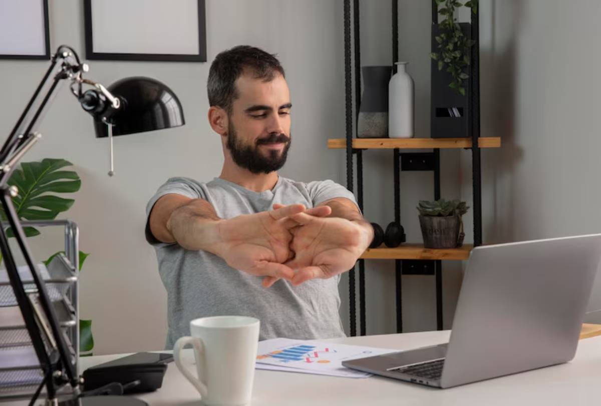 A person stretches their arms while seated at a desk cluttered with a laptop, a mug, and charts, in a cozy home office setting. 