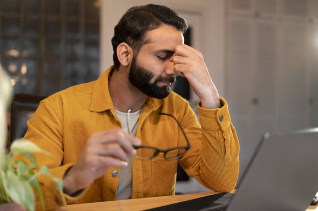 A man holds his eyeglasses in one hand while looking intently at a laptop screen, which is positioned on a desk in an indoor setting. The background features a wall, and the man is dressed casually.