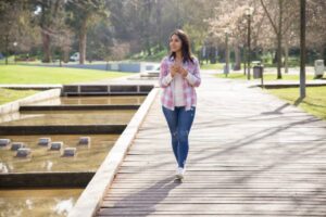 A woman walking on a wooden walkway surrounded by trees and grass, wearing jeans and footwear.