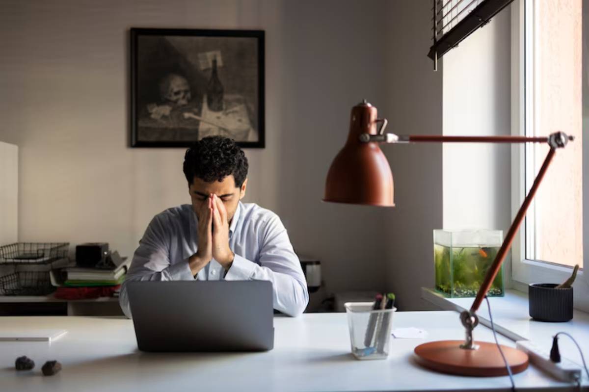 A person sits at a desk with a laptop, hands clasped, in a well-lit room featuring a desk lamp and a small fish tank. 
