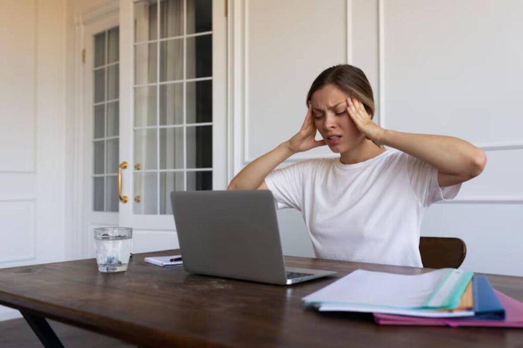 A person sitting at a wooden desk with a laptop, covering their ears, surrounded by papers and a glass of water.