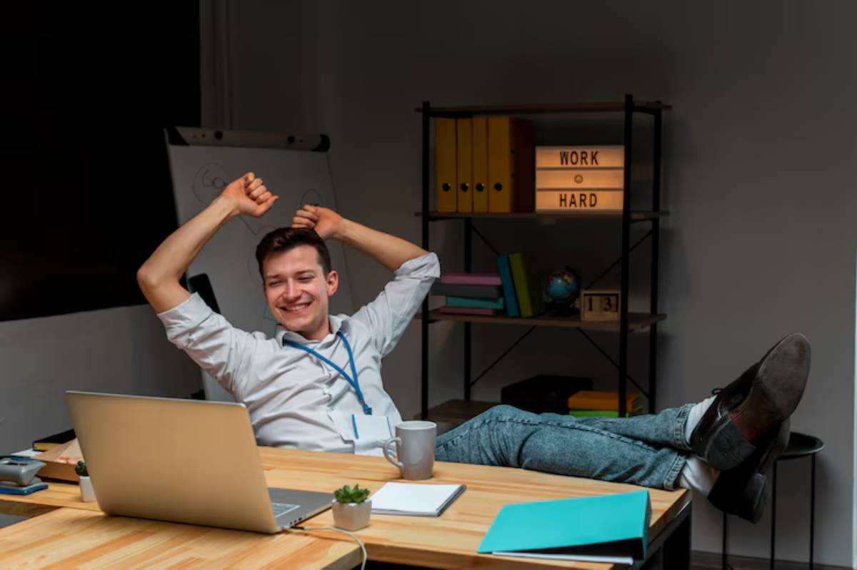  A relaxed individual sits at a cluttered desk, feet up, with a laptop, coffee cup, and motivational decor in a modern office space.