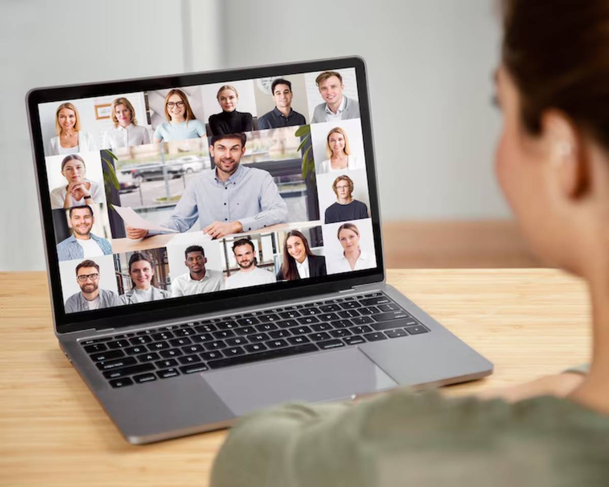 A person sitting at a desk looking at a laptop screen displaying multiple blurred faces in a video call setting. 