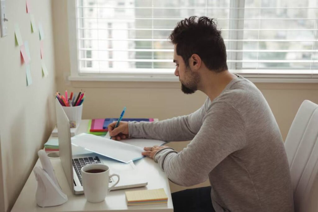 A person writing notes at a desk, with a laptop, coffee cup, and colorful stationery, bathed in natural light from a window.