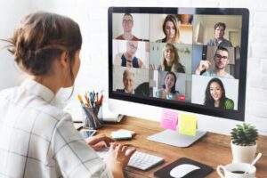 A person sits at a desk in front of a computer screen displaying a video call with multiple participants, surrounded by office supplies.