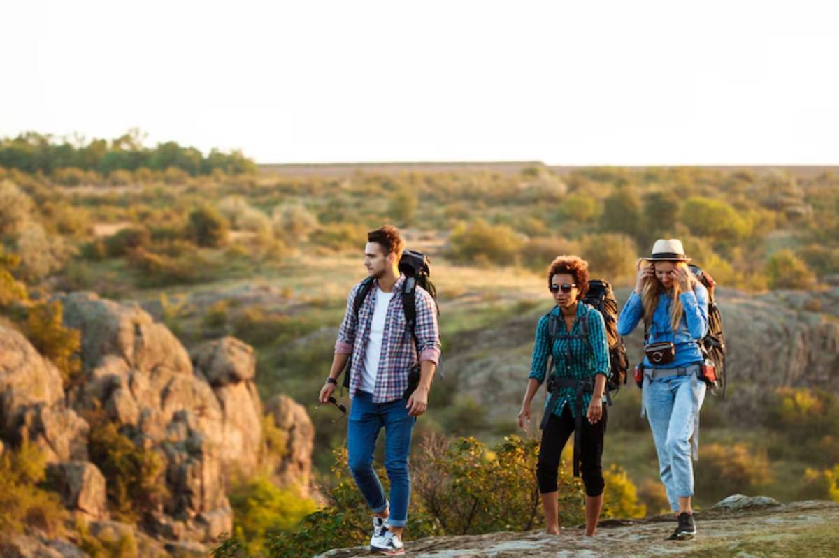 A group of people walking on a rocky surface outdoors, surrounded by grass and under a clear sky. They are wearing casual clothing including jeans and footwear appropriate for hiking, with a scenic mountain backdrop in the fall season. 
