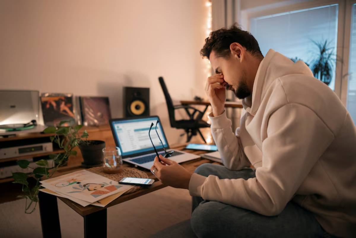  A person in a cozy room sits at a wooden table, surrounded by a laptop, smartphone, and art, deep in thought.