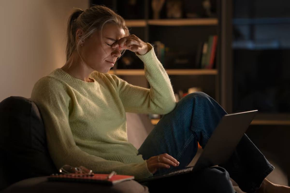 A person sits on a couch, wearing a light sweater and jeans, working on a laptop with a thoughtful expression in a dimly lit room. 