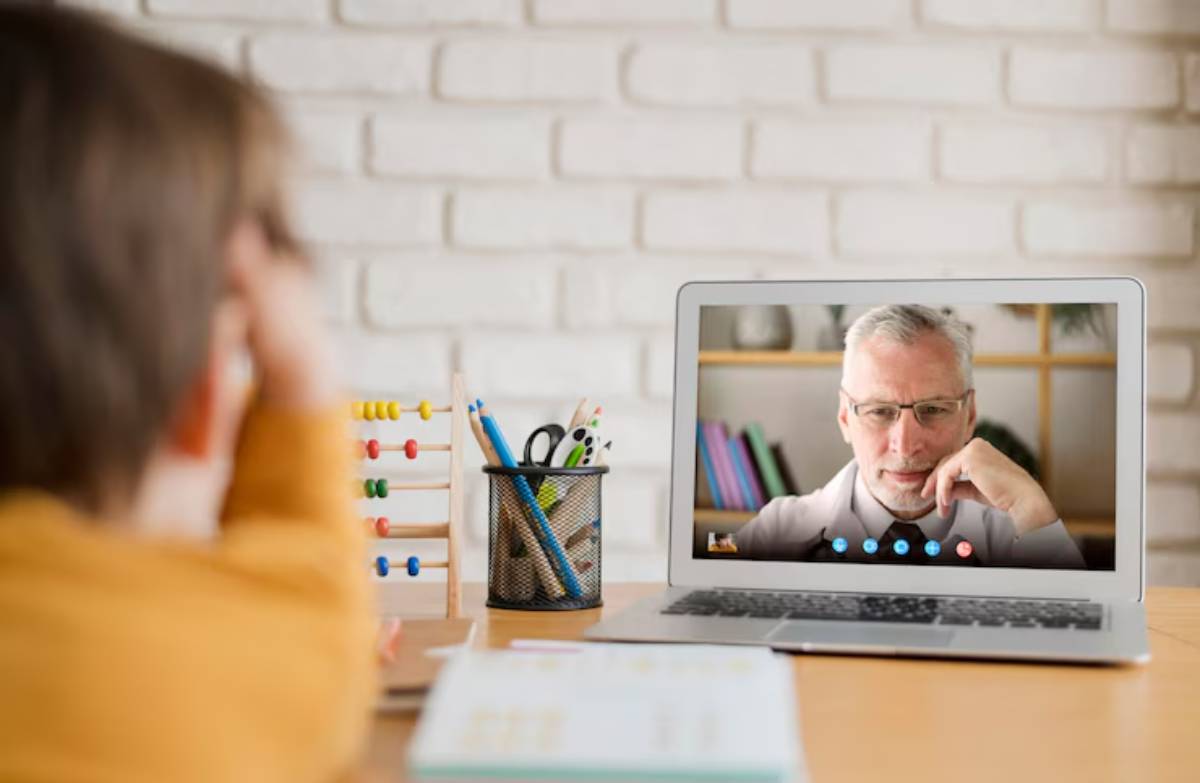 A child in a yellow shirt watches a video call on a laptop, with colorful stationery and an abacus visible on the table. 