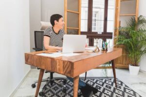 A person sits at a wooden desk with a laptop, surrounded by plants and a window, in a modern, well-lit office space.