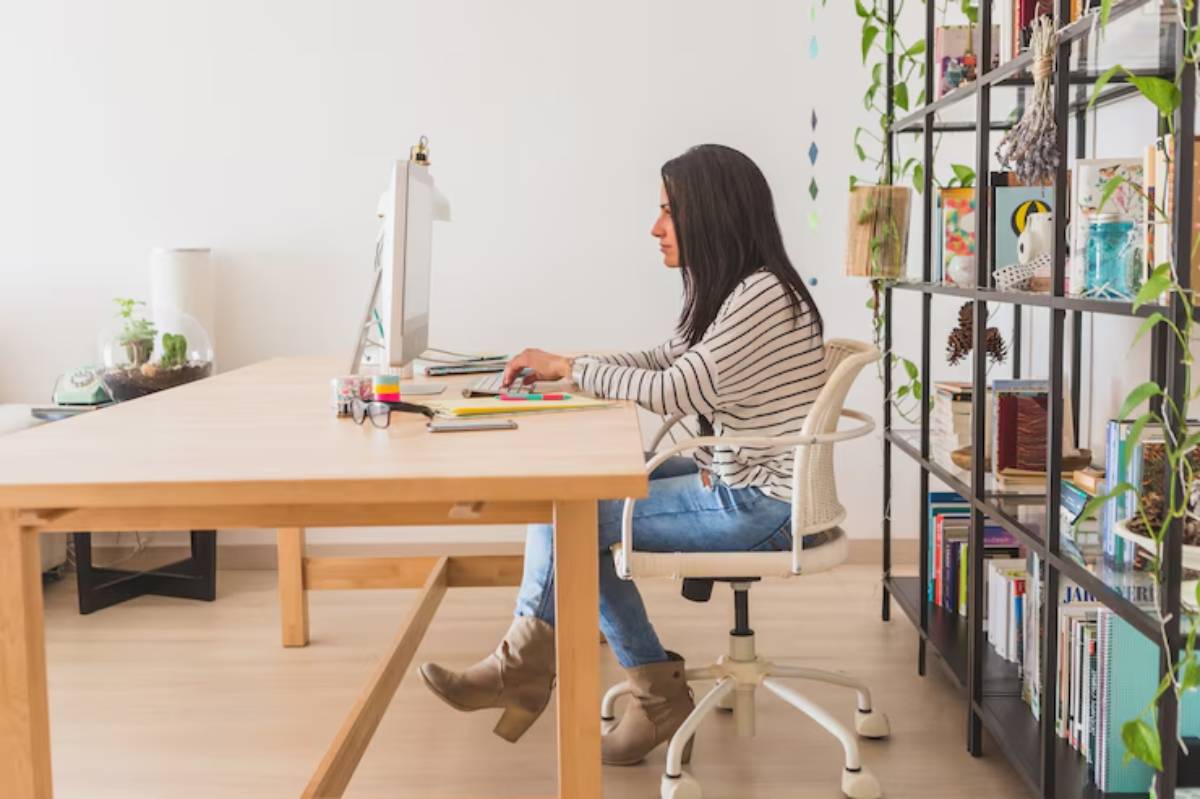 A person sits at a wooden desk, focused on a computer, surrounded by bookshelves and greenery in a bright, minimalist workspace. 