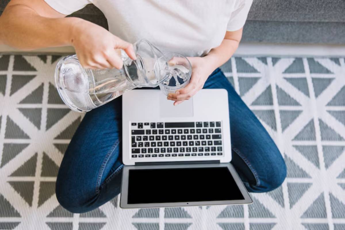 A person in jeans pours water from a glass pitcher into a cup, sitting cross-legged beside a laptop on a patterned rug. 
