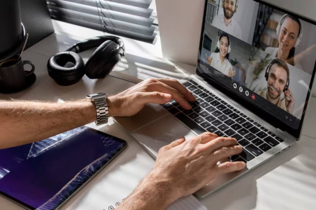 A person typing on a laptop with a video call interface, surrounded by headphones and a notepad on a bright desk.
