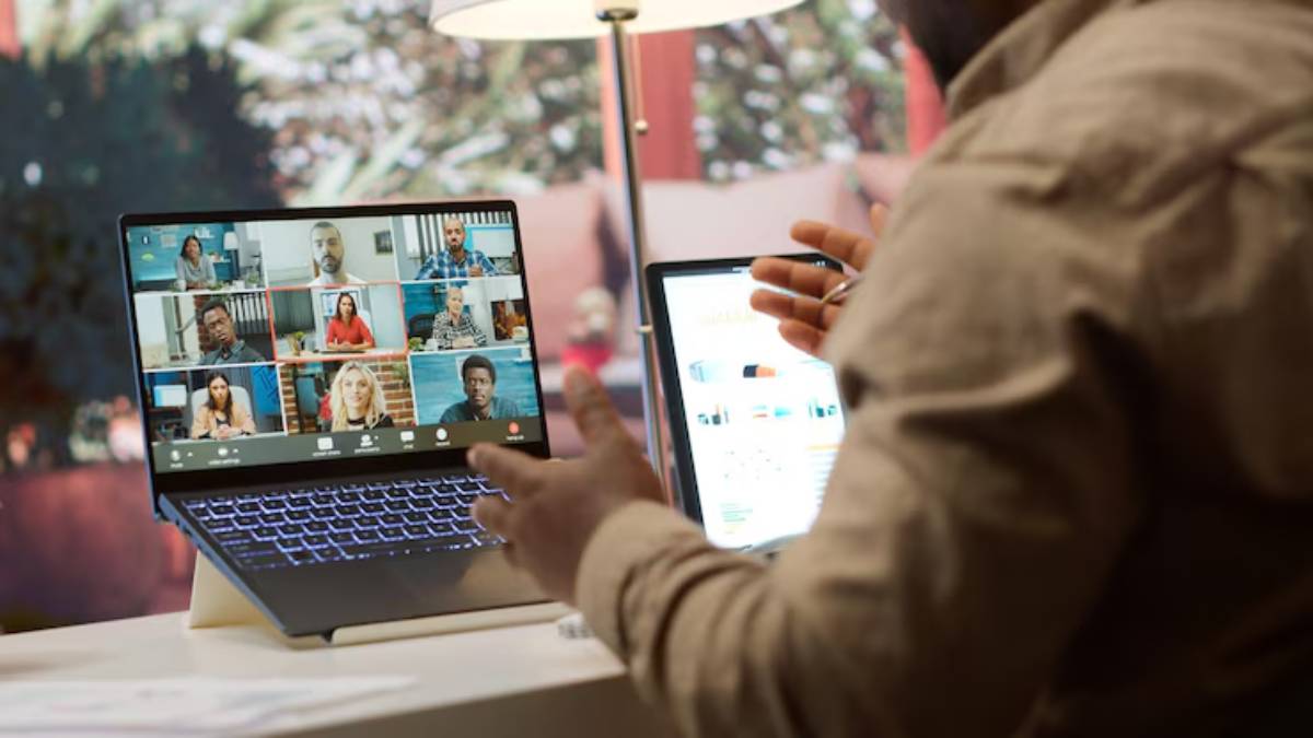 A person participates in a virtual meeting on a laptop, gesturing with a pen in hand, while a tablet displays additional documents. 