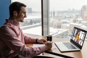 A person wearing wireless earbuds sits at a desk, taking notes while attending a video conference on a laptop, with a snowy city view.