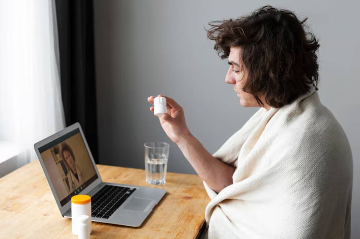 A person wrapped in a blanket holds a pill bottle and sits in front of a laptop, engaged in a video consultation, with a glass of water nearby. 