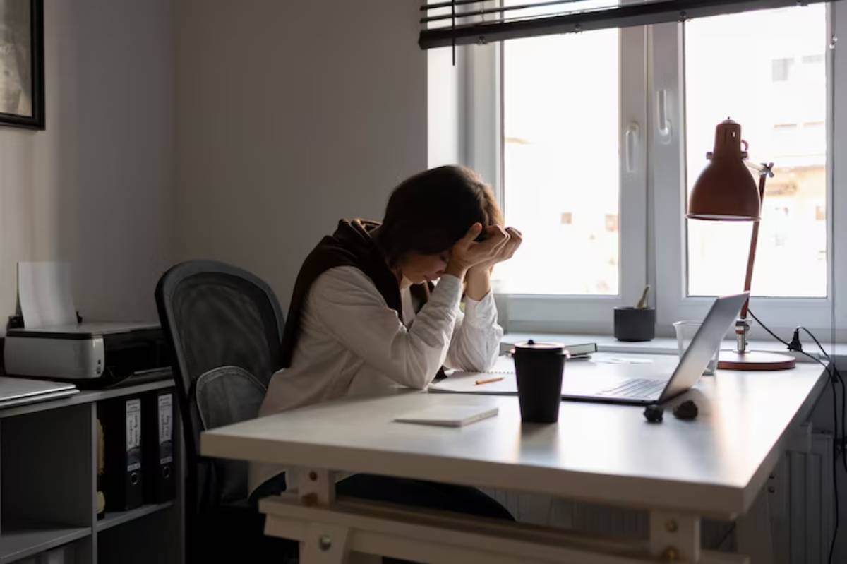 A person sits at a desk, resting their head on their hands in a sunlit office with a laptop and a coffee cup nearby. 
