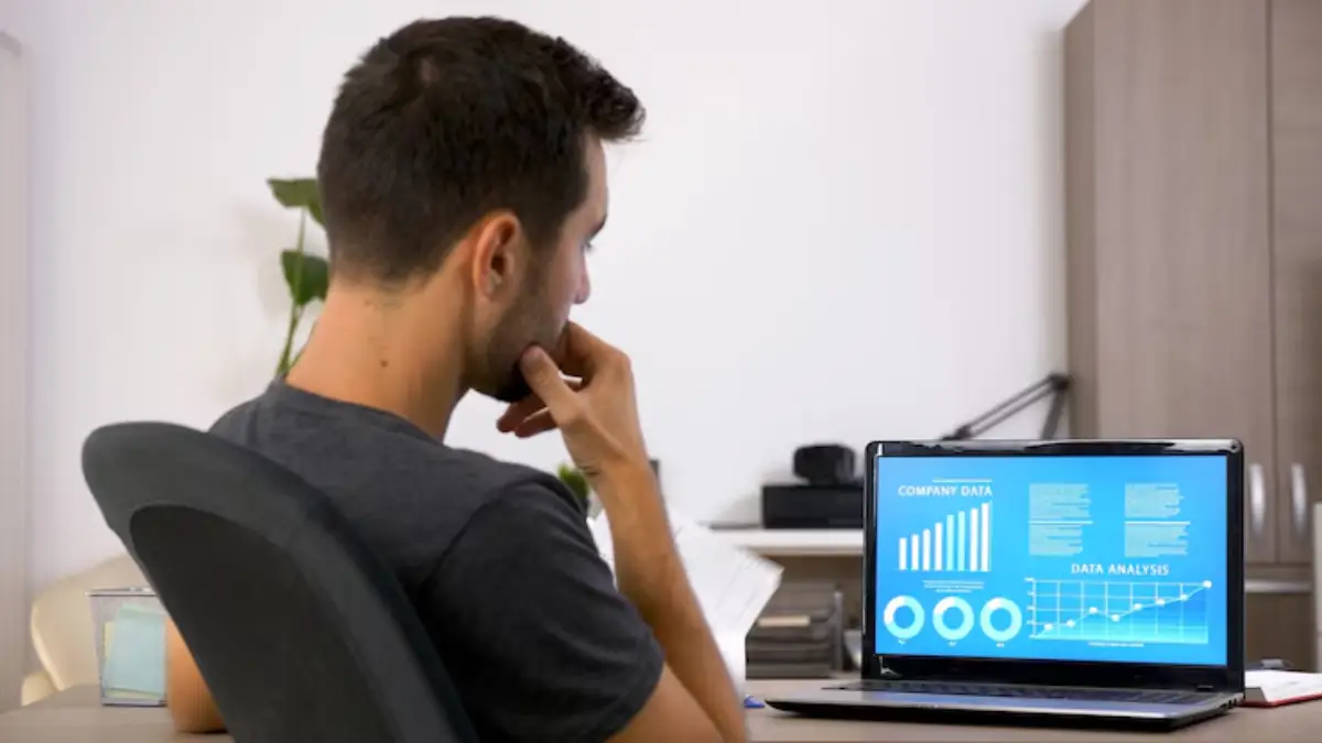 A person seated at a desk, looking thoughtfully at a laptop displaying data analysis graphs and charts. 