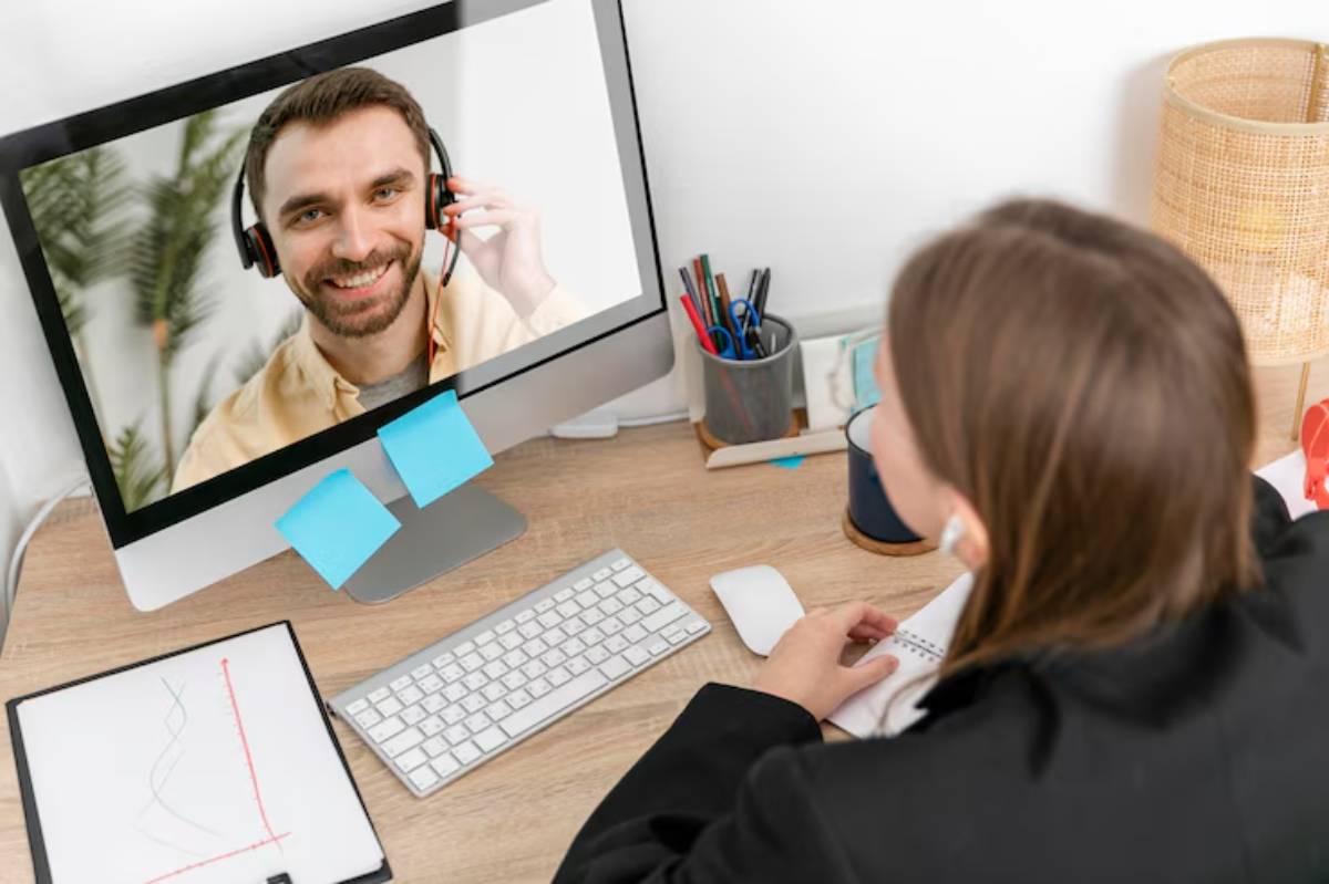  A woman in a black jacket sits at a desk, engaged in a video call on a computer, surrounded by office supplies and a warm lamp.