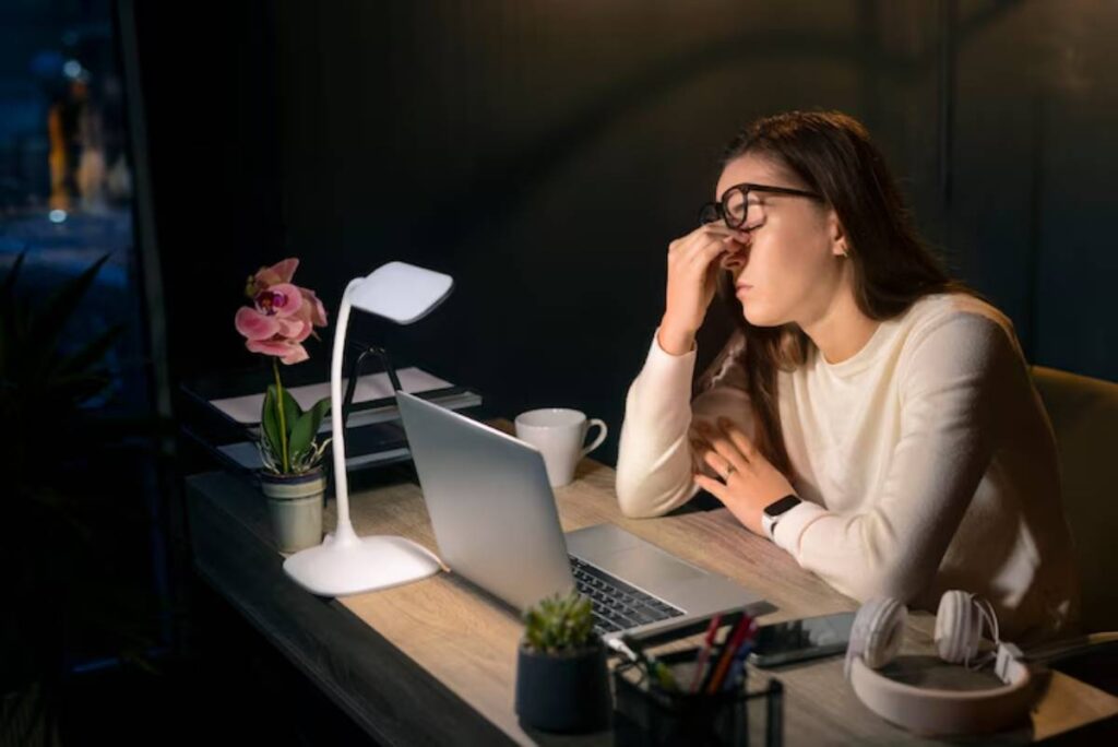 A person sits at a desk with a laptop, a plant, a cup, and stationery, focused on work in a dimly lit room.