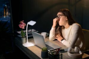 A person sits at a desk with a laptop, a plant, a cup, and stationery, focused on work in a dimly lit room.