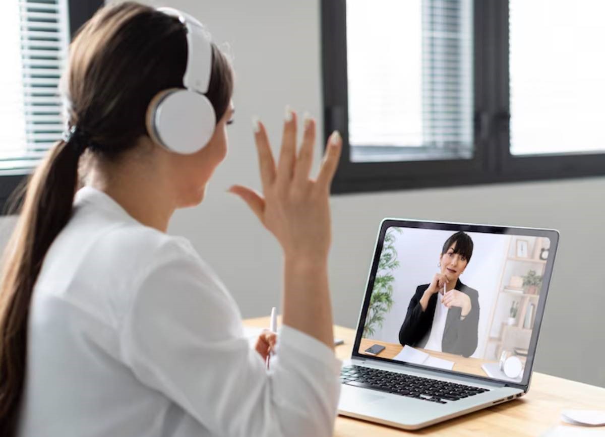 A woman wearing headphones gestures while engaged in a video call, sitting at a wooden desk with a laptop and notepad. 