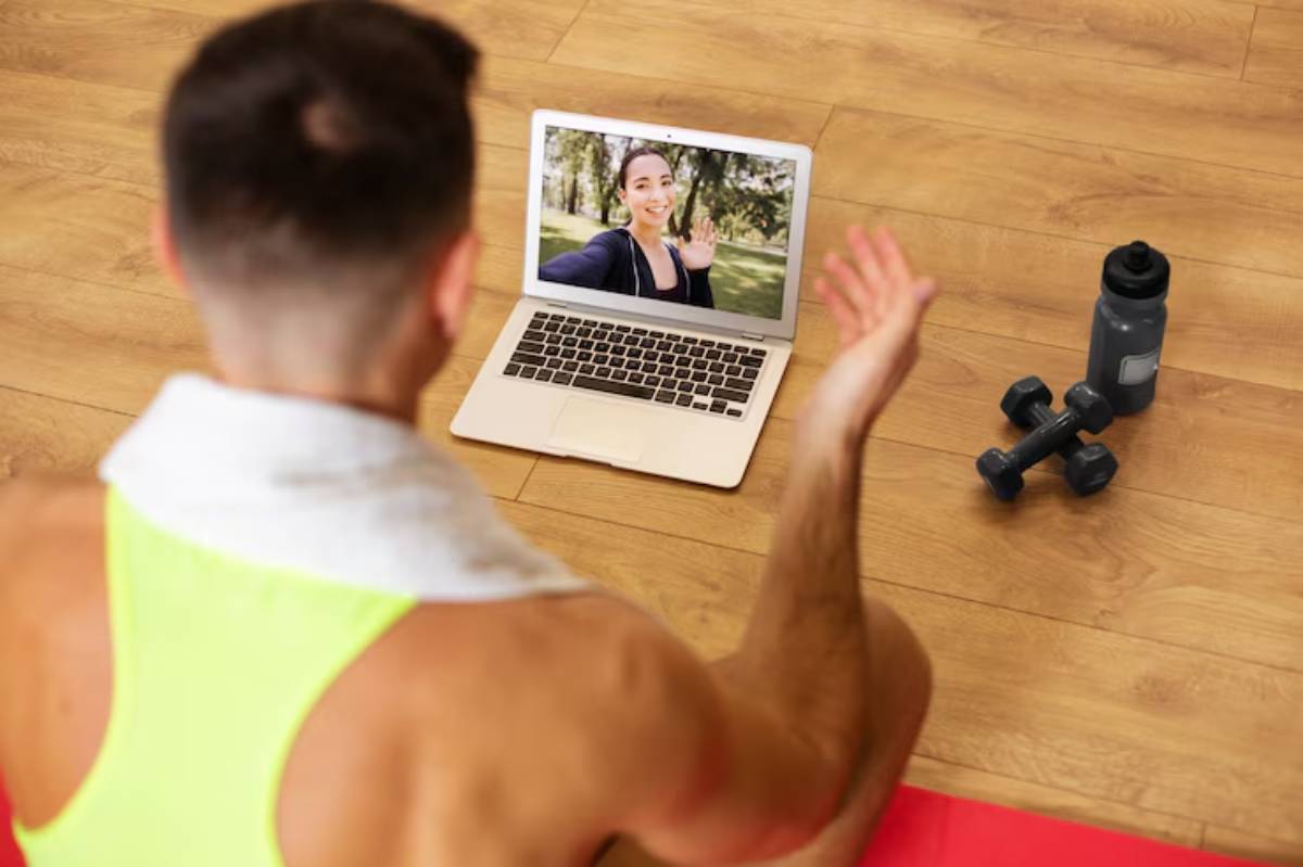A person in a bright tank top interacts with a laptop on a mat, with dumbbells and a water bottle nearby. 