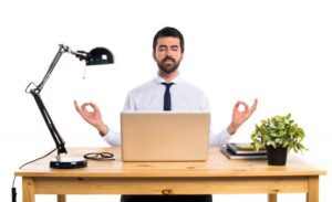 A man in a white shirt and tie sits at a desk, meditating with hands in a gesture of peace, a laptop, lamp, and plant nearby.