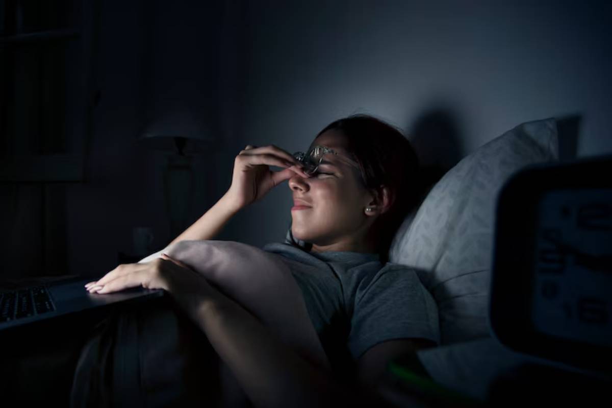  A woman lying in bed with her hand on her nose, set against a dark backdrop.
